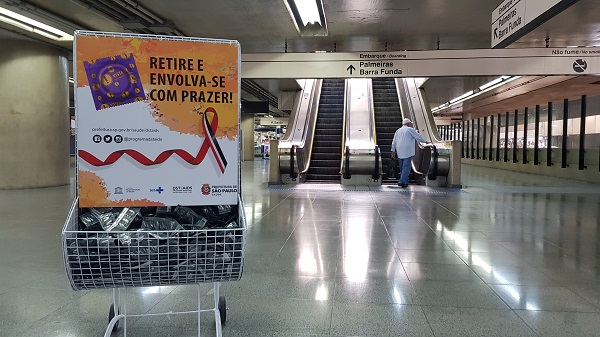 #PraCegoVer: Display de camisinha está posicionado no cano esquerdo da foto. No fundo, um munícipe sobe as escadas rolantes da estação.